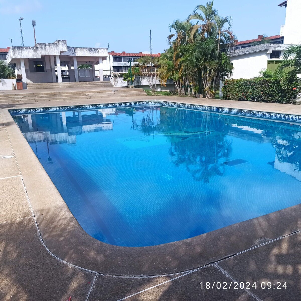 A large swimming pool is set within a well-maintained residential area, surrounded by tropical plants and palm trees. The water is clear and reflects the sky, while lounge chairs are positioned around the poolside for relaxation.