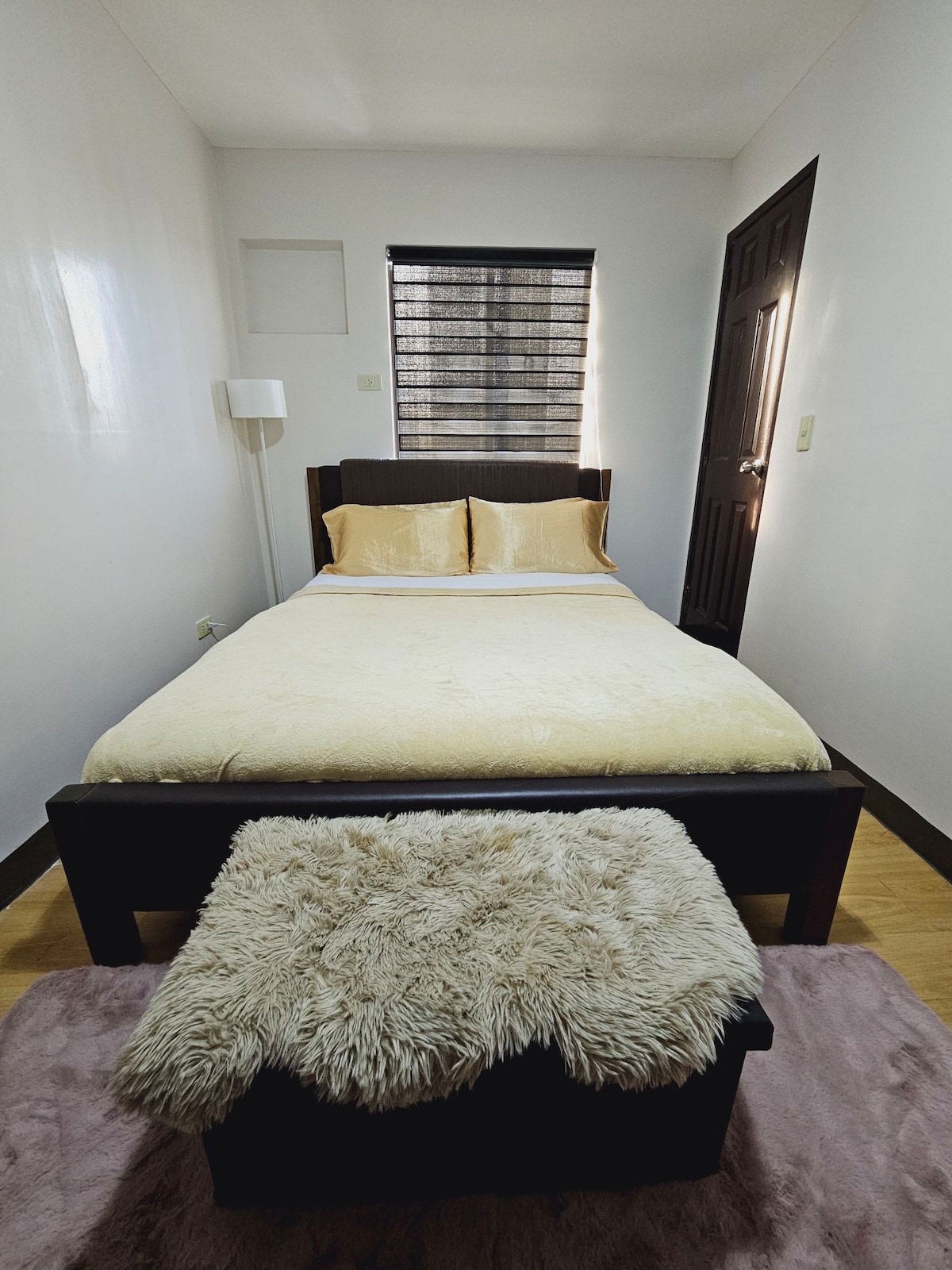 A bright sleeping area is presented, featuring a spacious bed with beige linens and two gold accent pillows. A plush, light-colored stool is positioned at the foot of the bed. Natural light filters through a window adorned with black blinds, complementing the room's minimalist design.