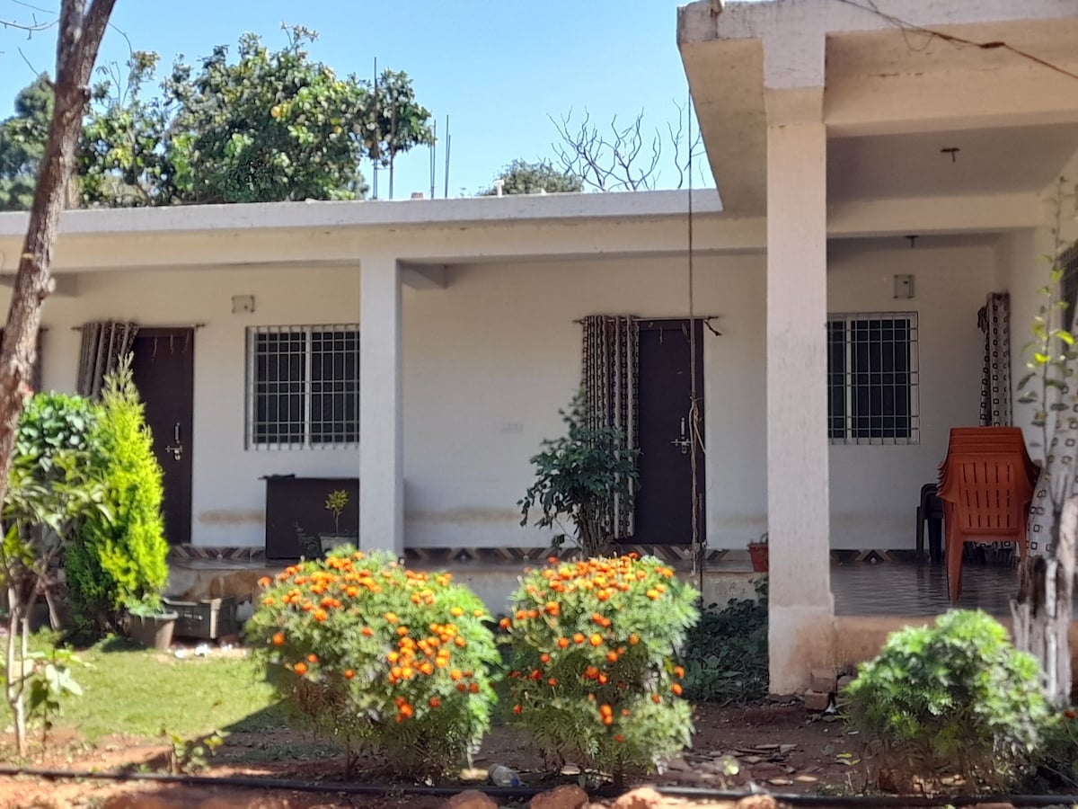 A welcoming exterior view of the property featuring well-maintained garden beds with vibrant orange and green plants. Two separate entrances are visible, framed by simple windows with closed curtains. A small seating area is positioned next to the vibrant greenery, inviting relaxation.