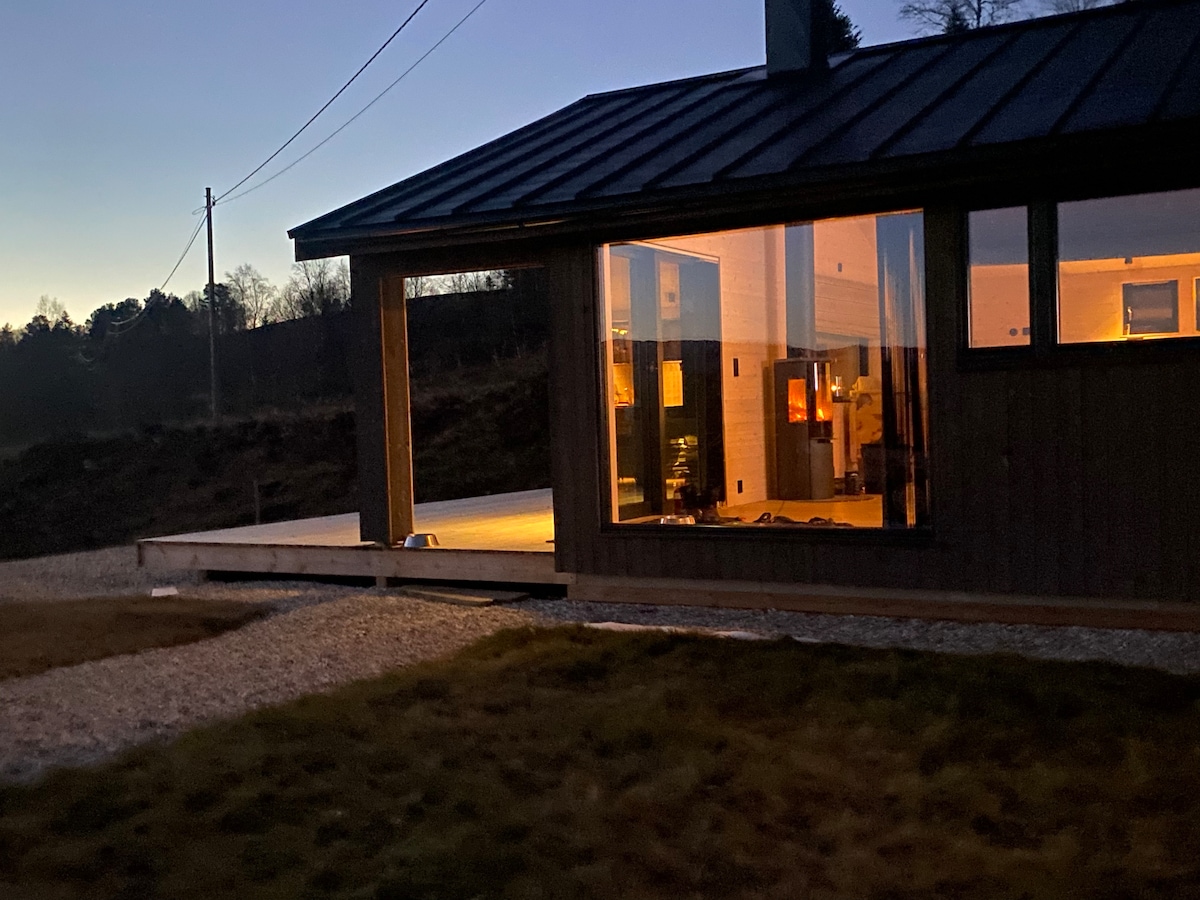 The exterior of the cabin is shown during twilight, with warm light emanating from large windows. A wooden deck extends towards the grassy area, framed by gravel pathways. Surrounding trees and a serene landscape create a calming backdrop, enhancing the peaceful atmosphere.