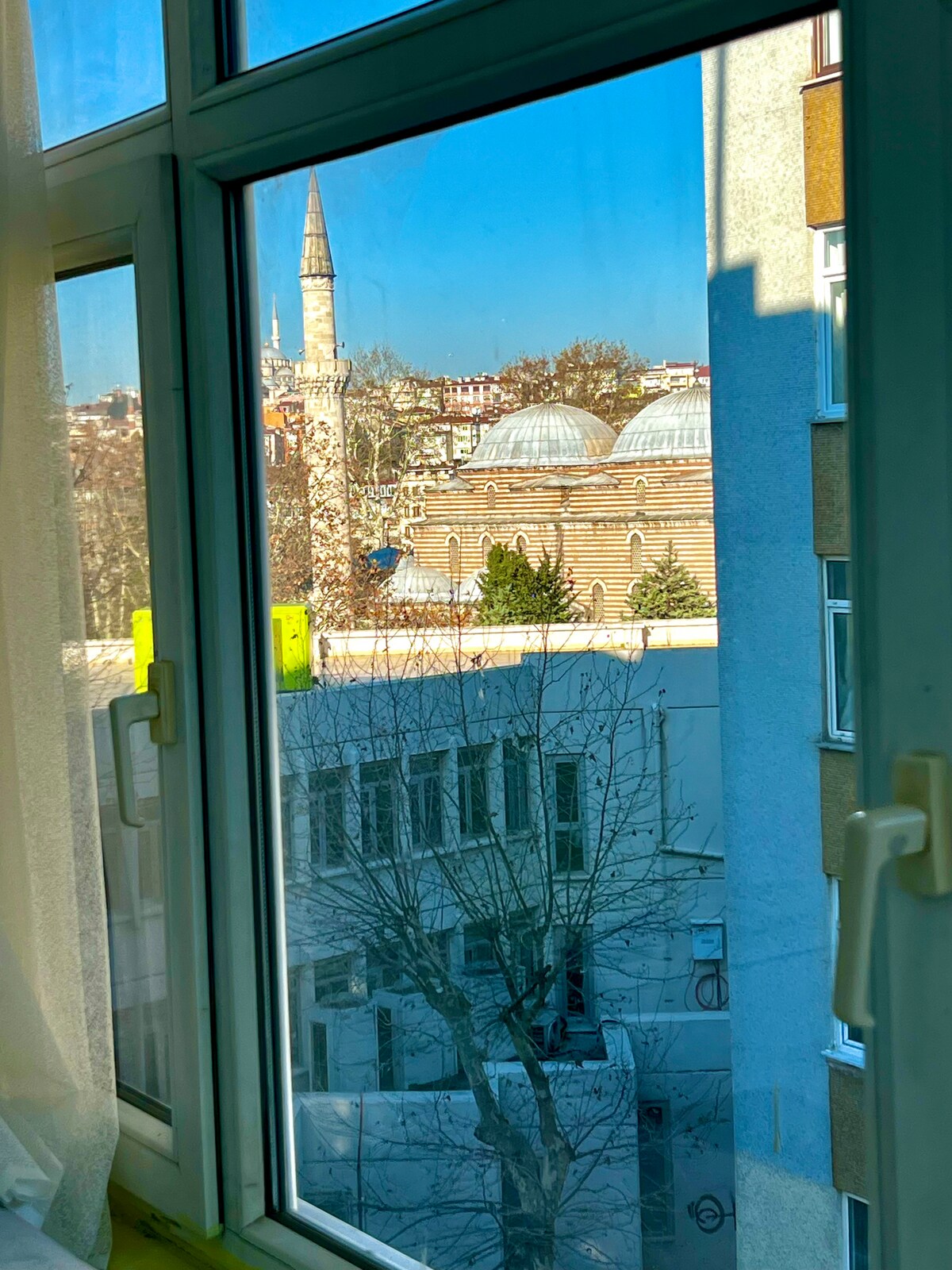 A view from the window reveals an inviting scene of a historic mosque with a tall minaret. The architecture showcases a blend of stone and domes, while the tree outside adds a touch of nature to the urban setting. Bright light filters through the window.