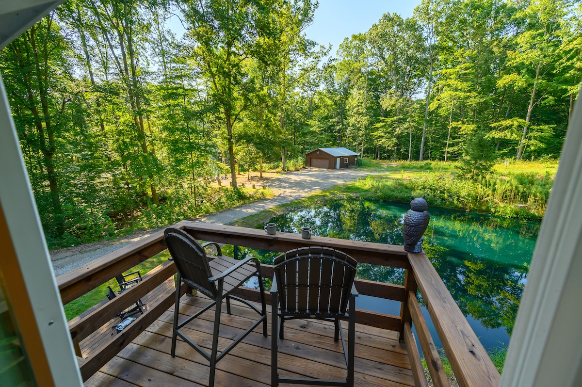 A scenic view of a tranquil pond is presented from a wooden deck, featuring two outdoor chairs. Lush greenery surrounds the water, with a glimpse of a storage building visible in the background, enhancing the natural setting.