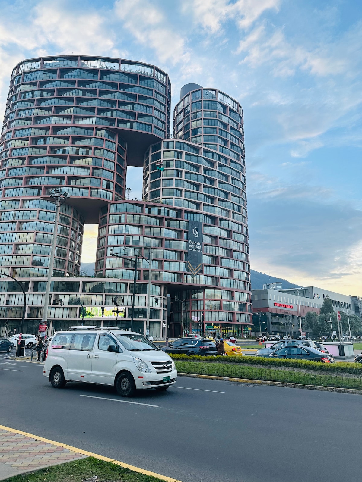 A modern architectural building features a unique design with a series of interconnected curves and glass facades. The structure showcases varied heights and shapes, complemented by surrounding urban scenery, with vehicles and pedestrians visible on the nearby road.