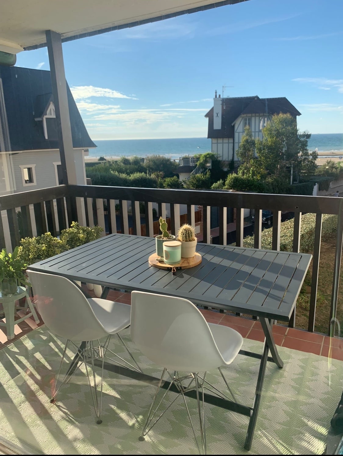 A balcony features a rectangular table surrounded by two white chairs, offering a view of the sea and nearby buildings. Potted plants, including a succulent, are displayed at the center of the table, with sunlight illuminating the serene outdoor space.