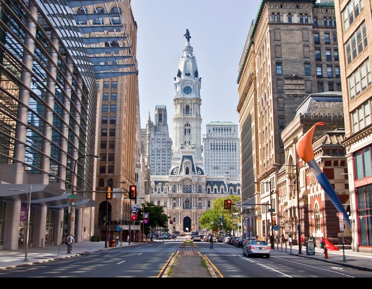 The image captures a bustling city street leading to a grand clock tower at City Hall, characterized by its intricate architectural design. Surrounding buildings exhibit diverse styles, with modern glass structures juxtaposed against classic facades. Traffic and pedestrians add to the vibrant urban atmosphere.