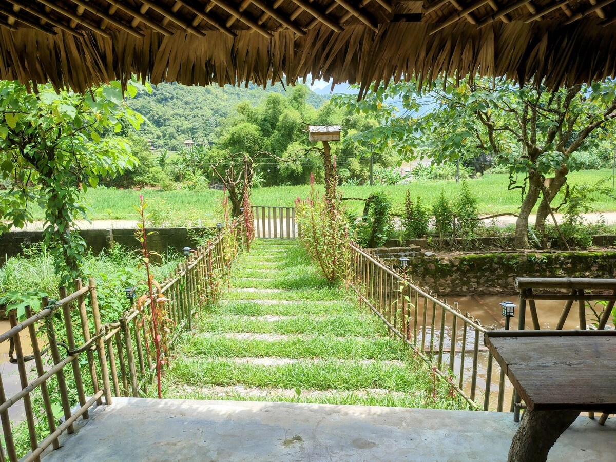 A pathway lined with vibrant green grass and bordered by plants leads to a serene outdoor space. The view showcases open fields and a backdrop of lush hills, framed by a rustic wooden structure with a thatched roof.