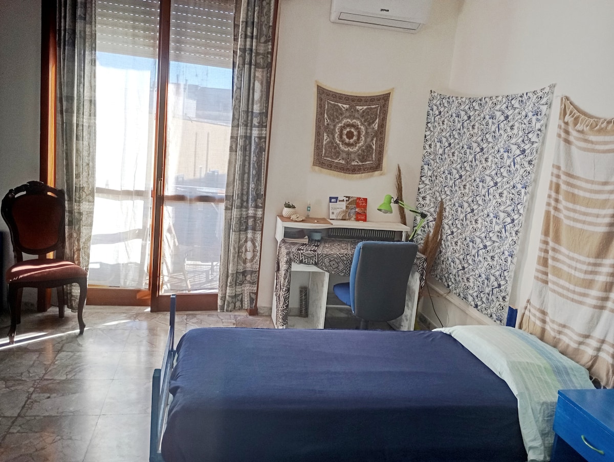 A cozy shared room is equipped with two single beds dressed in blue linens. Natural light streams in through large windows with sheer curtains, illuminating a small desk and chair. A decorative wall tapestry and potted plants add a touch of warmth to the space.
