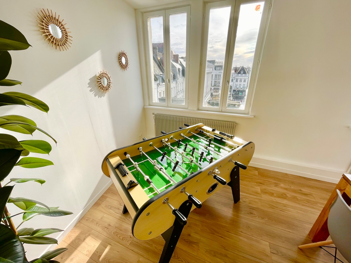 A foosball table is situated in a well-lit corner, with sunlight streaming through large windows. The green playing surface is surrounded by a minimalistic decor, highlighted by round mirrors on the wall and natural wooden flooring, creating a pleasant environment for leisure activities.