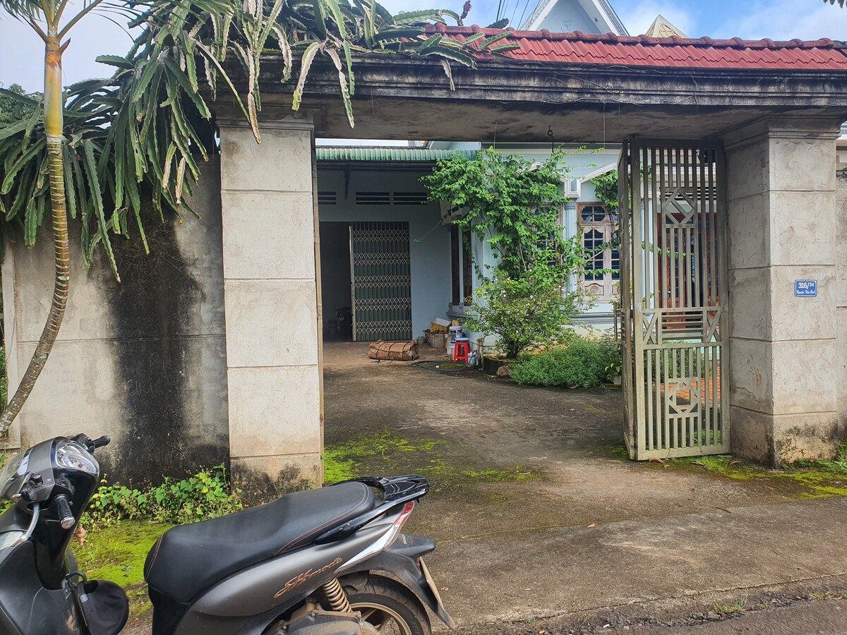 An entrance to a spacious outdoor area is framed by stone pillars, leading to a welcoming pathway. Lush greenery and potted plants are visible, adding a natural touch. A motorcycle rests on the left side, with a clear sky overhead and well-maintained surroundings.