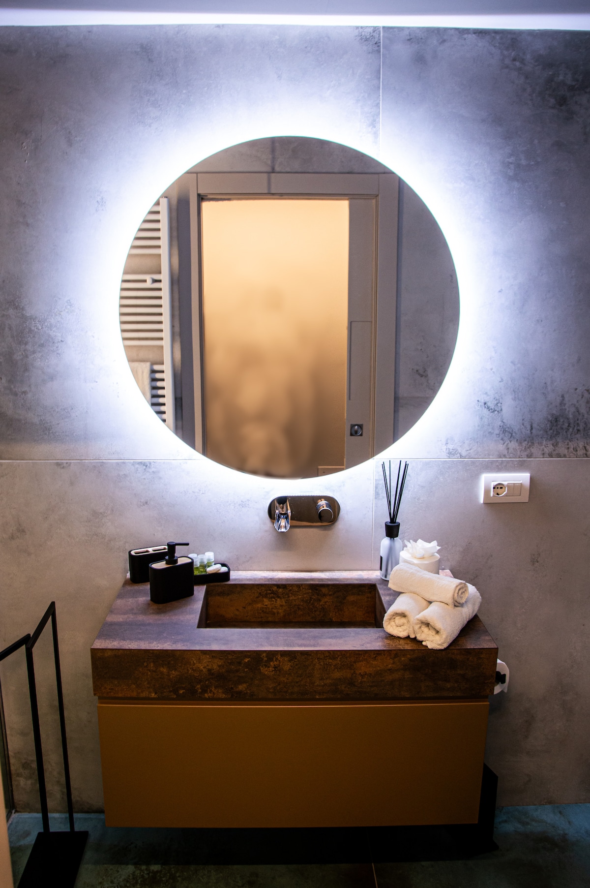 The elegant bathroom features a round illuminated mirror and a stylish sink with a modern finish. Soft towels are neatly arranged beside natural toiletries, while a bathroom accessory set adds a touch of sophistication. The muted tones create a calming atmosphere.