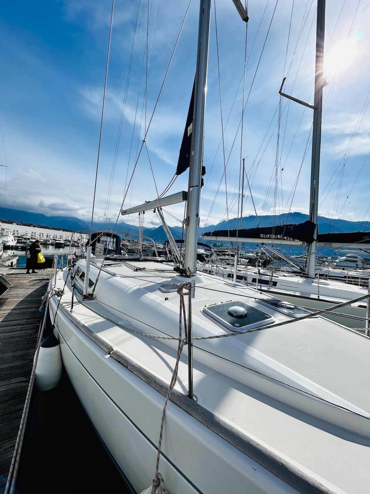 A sleek sailing yacht is docked beside a marina, showcasing its bright white exterior. The mast and rigging rise prominently against a clear blue sky, with several nearby boats visible in the background. Sunlight glints off the deck, highlighting the yacht's well-maintained features.