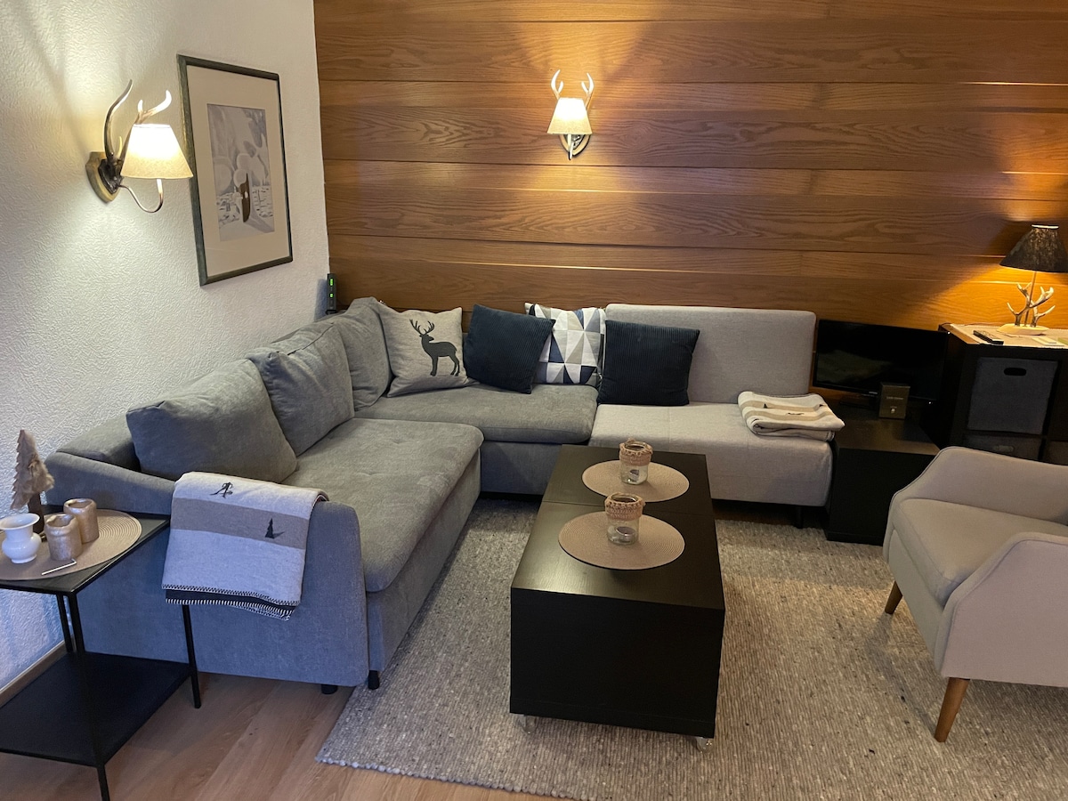 A spacious living area features a large, L-shaped gray sofa adorned with decorative cushions. A small black coffee table sits in front, complemented by a cozy armchair. Soft lighting is provided by wall sconces, enhancing the overall warmth of the wood-paneled walls.