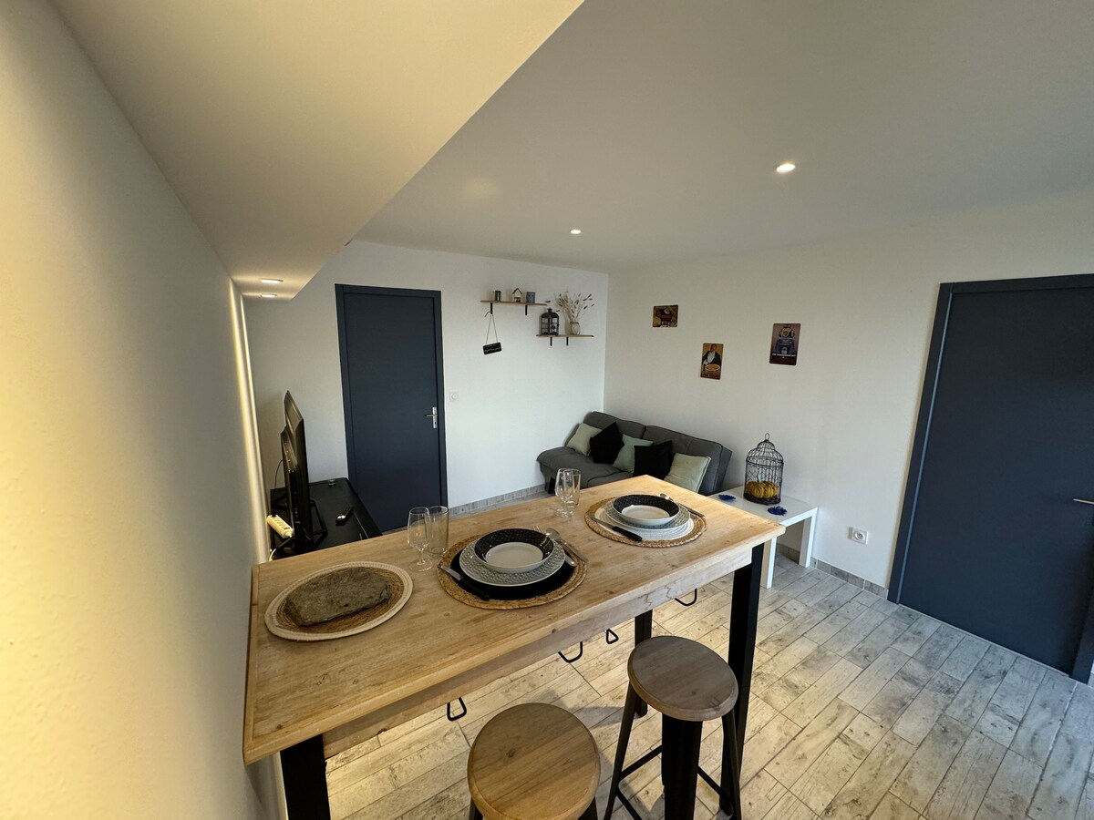 The living space features a wooden dining table set for four, with three stools positioned nearby. A gray sofa is placed against the wall, facing a television. Light-colored walls and flooring enhance the room's brightness and openness.