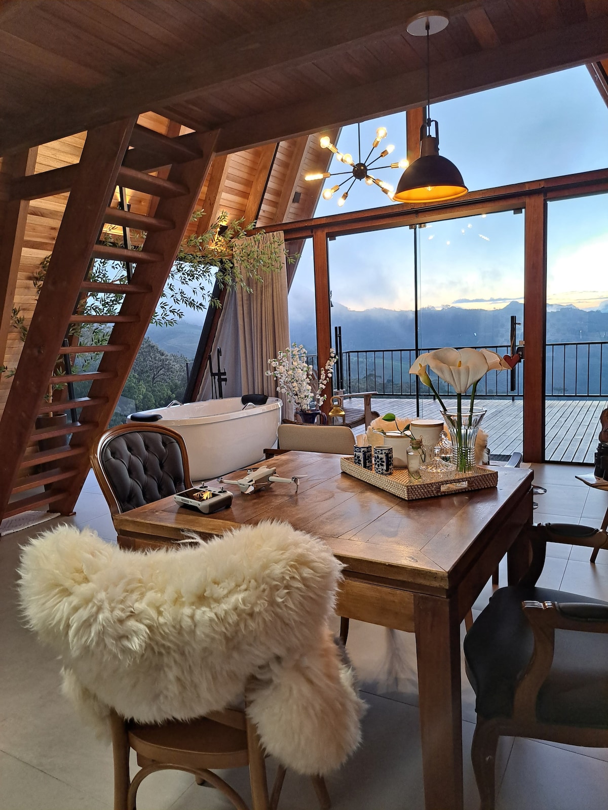 A dining area is presented with a wooden table adorned with decorative items and a vase of flowers. A fur throw drapes over a chair, and large windows offer sweeping views of the surrounding mountains, inviting natural light into the space.