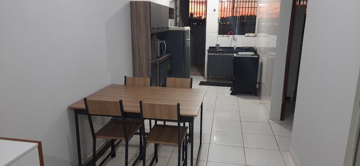 A modern kitchen and dining area is presented, featuring a rectangular wooden table with four chairs. The kitchen includes dark cabinetry, a microwave, and ample counter space, with a large window allowing natural light to fill the room.