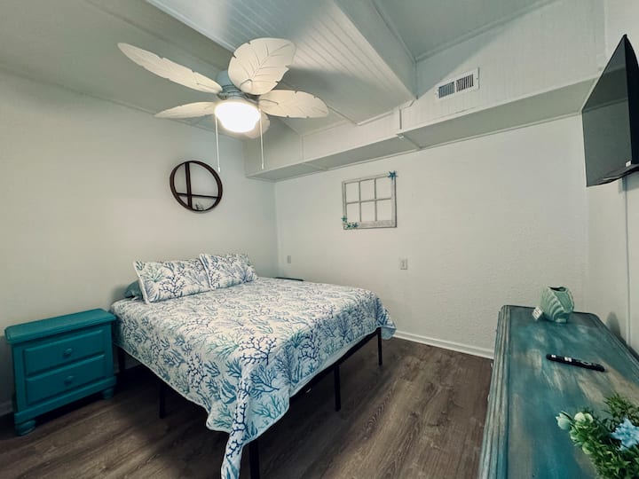 Coastal bedroom with plush bed, TV, leaf fan, and beachy decor.