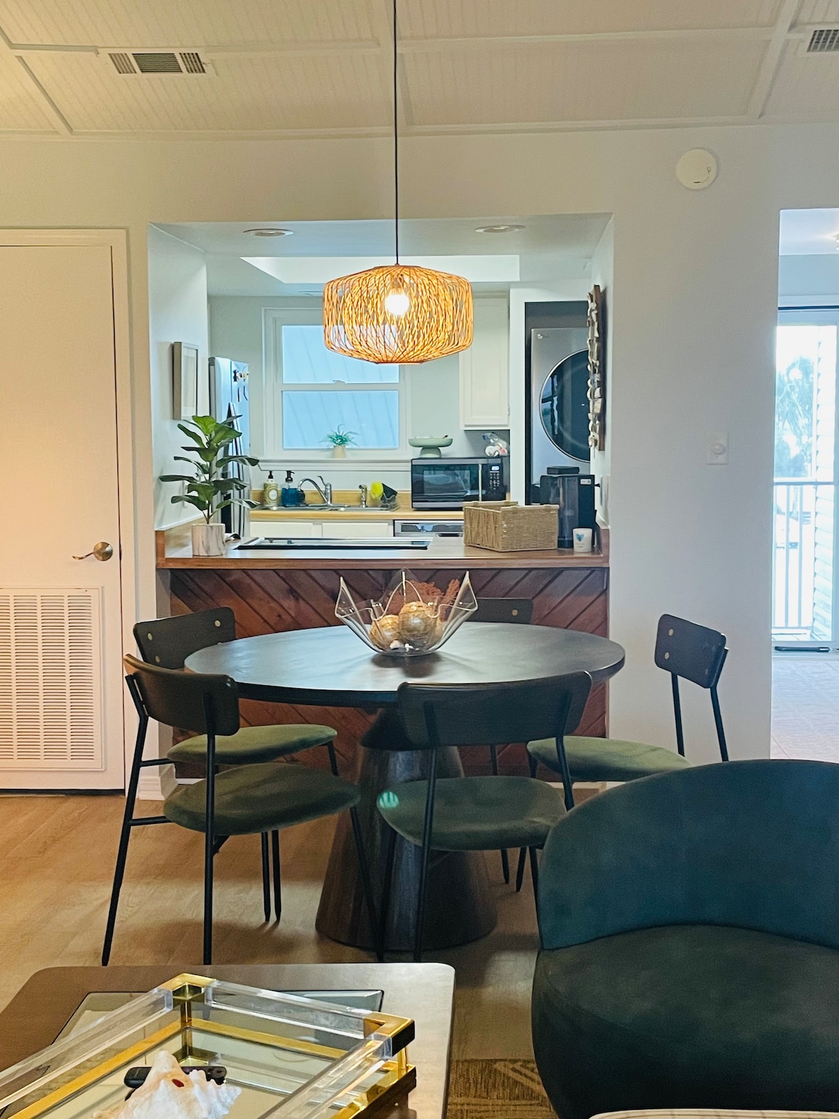 A modern dining area features a circular wooden table surrounded by four sleek chairs. A decorative bowl sits in the center of the table. Soft lighting is provided by a woven pendant above, while the kitchen area can be seen in the background.
