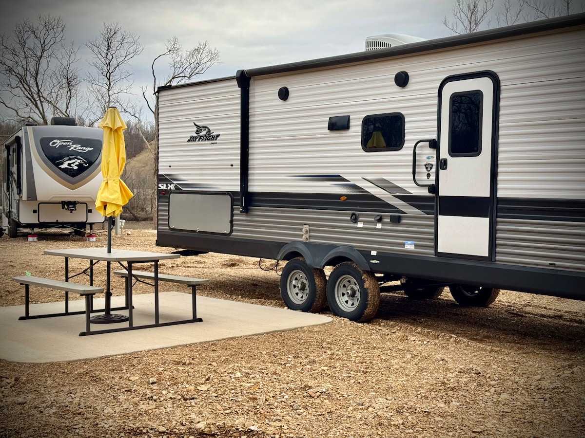 A Jayco camper is featured, showcasing a sleek exterior design. A picnic table with an umbrella is situated on a concrete pad beside the camper, set against a backdrop of gravel and bare trees. Another camper is visible in the background.