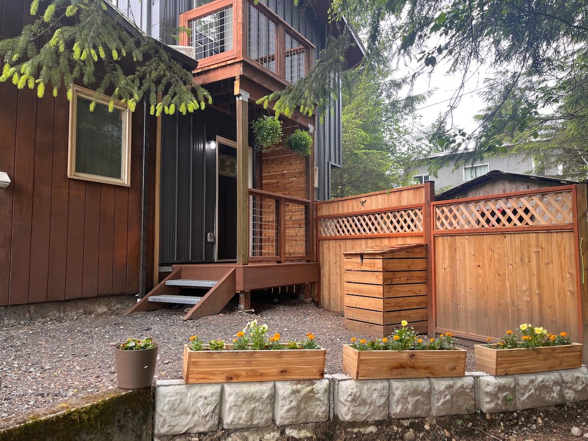 The exterior of the attached apartment showcases a welcoming entrance with wooden decking and a private fenced area. Planters with colorful flowers line the gravel path, and a wooden storage box is visible beside a lattice fence, offering charm and privacy.