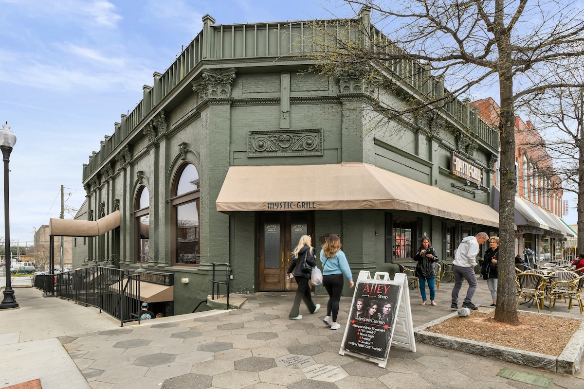 A historic building with dark green exterior paint features large arched windows and a canopy shading the entrance. Several pedestrians are seen walking by, along with outdoor seating areas. A chalkboard sign displays event information outside the venue.