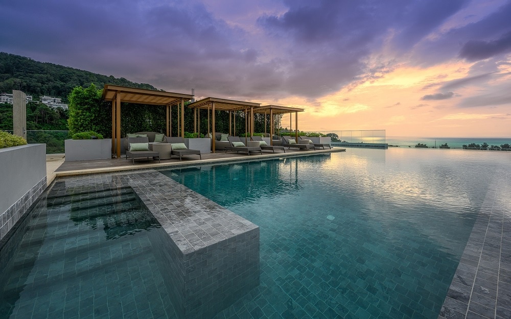A serene infinity pool stretches across the image, reflecting the colorful sky at sunset. Lounge seating is positioned under wooden cabanas, offering a relaxing space. Lush greenery borders the area, enhancing the outdoor environment.