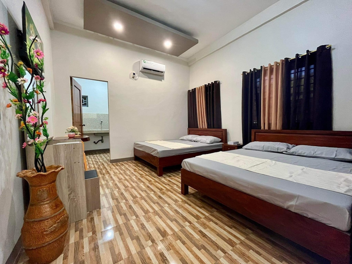 A spacious room features two wooden beds arranged with light-colored linens. A decorative vase filled with flowers stands to one side. Large windows are dressed with dark curtains, while an air conditioning unit is mounted above. A mirror and a bathroom door are visible in the background.