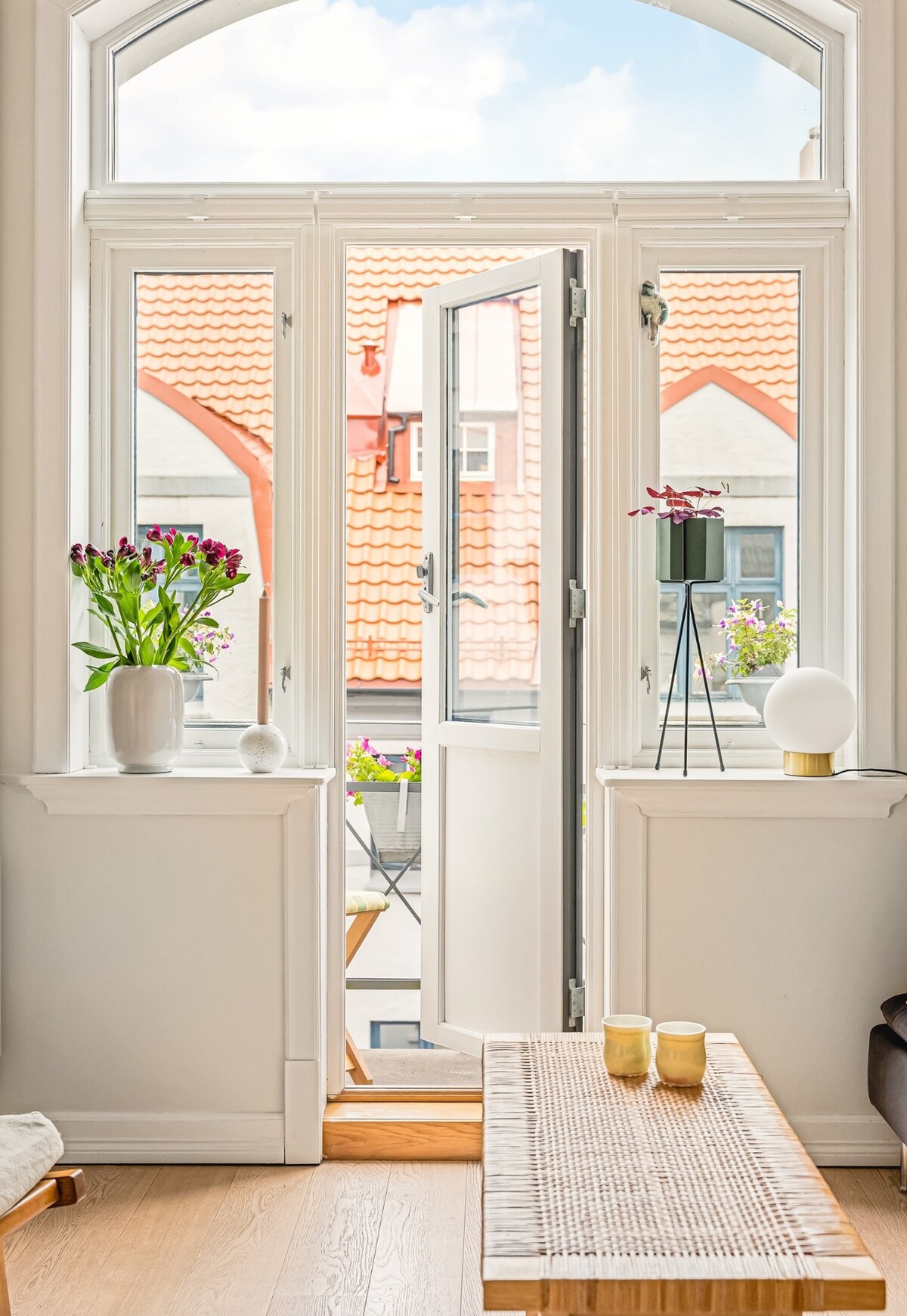 A bright room is showcased with wide-open doors leading to a balcony. Potted flowers add greenery, while a stylish table lamp and decorative items sit elegantly on a console. Natural light fills the space, highlighting the warm tones of the wooden flooring.