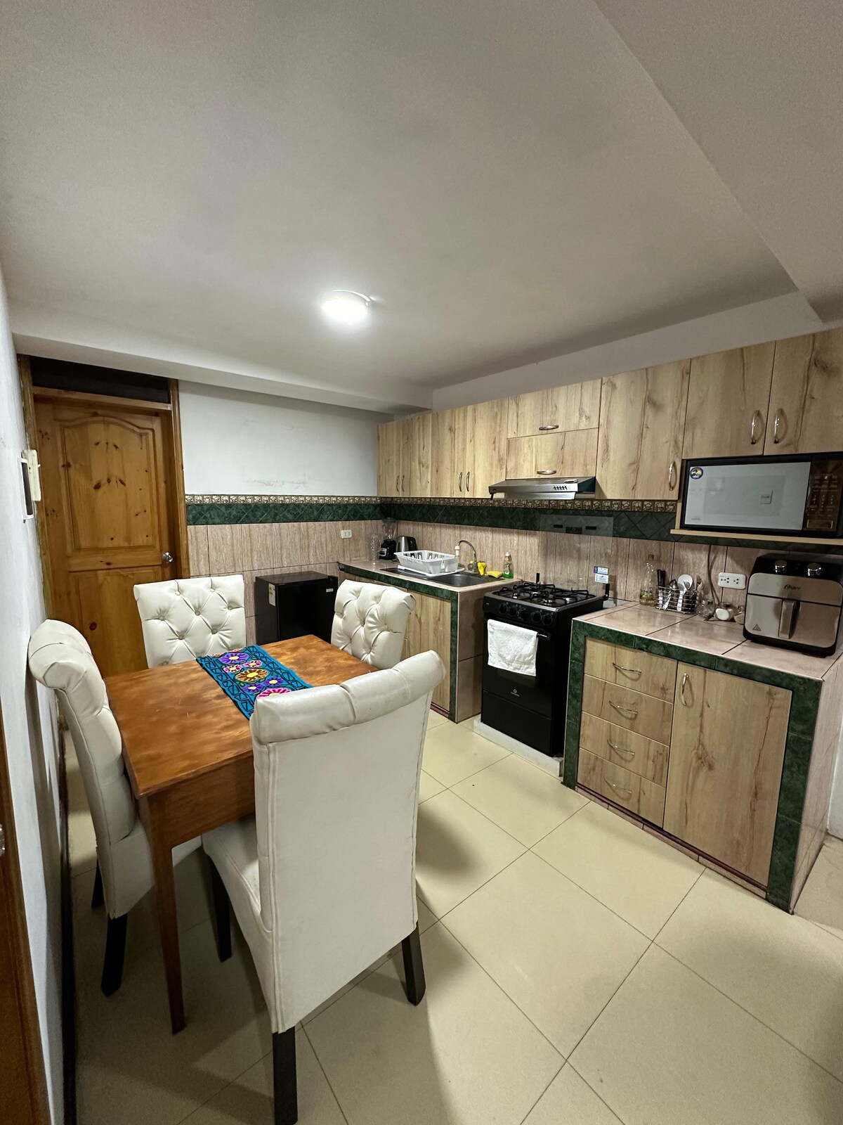 A well-equipped kitchen features wooden cabinetry with a blend of light and dark tones. A dining table with four upholstered chairs occupies the center, while a stove, microwave, and sink are arranged along the counter. Soft lighting enhances the functional layout.