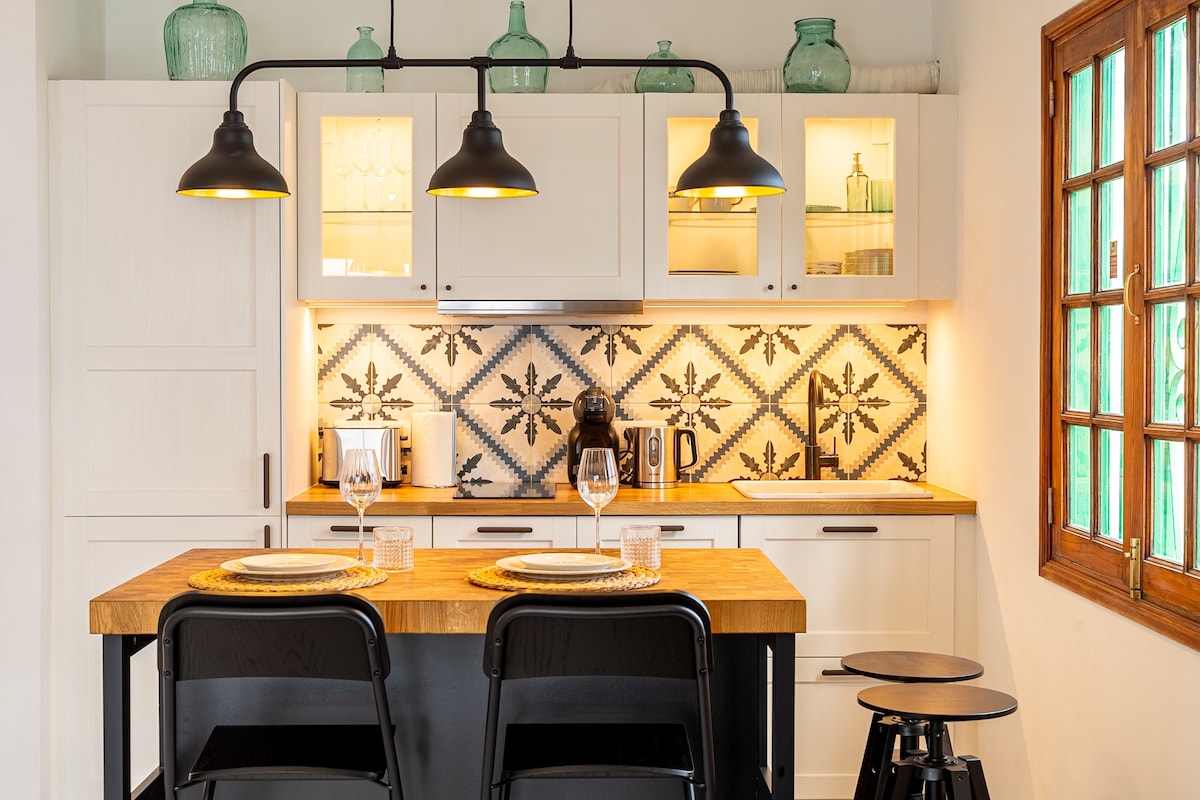 A modern kitchen area features a wooden countertop with two black chairs facing it. Above, pendant lights illuminate the space. The cabinetry displays glassware, while an intricately patterned backsplash adds a visual focal point. Natural light enters through a wooden window with multiple panes.