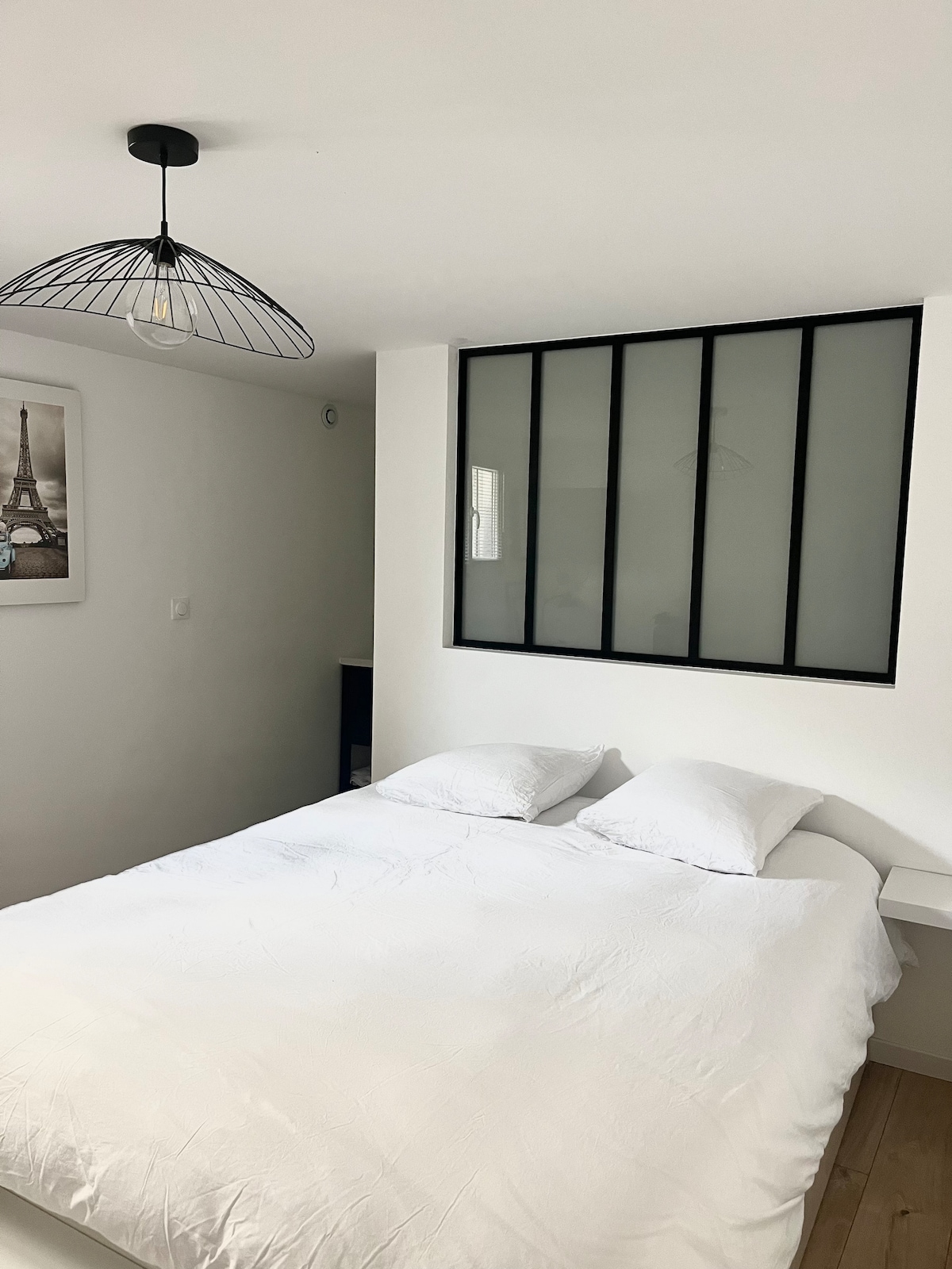 A spacious sleeping area features a large bed with a simple white duvet and two pillows. Natural light filters through a modern glass-paneled wall, complementing the minimalist design. A framed artwork of the Eiffel Tower is displayed on the adjacent wall.