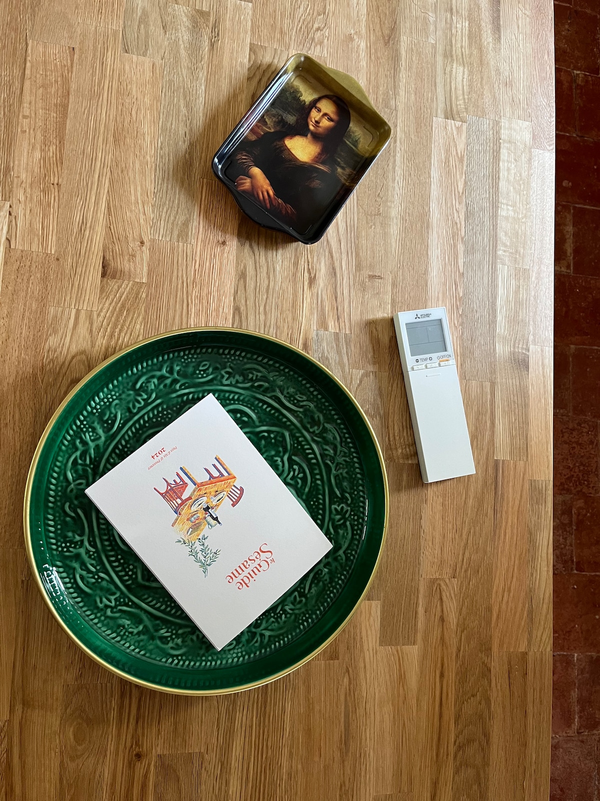 A wooden table showcases a green decorative tray, a floral-themed postcard, a rectangular metal tin with a lid, and a remote control. The arrangement features varying textures and colors, with natural light illuminating the surface.
