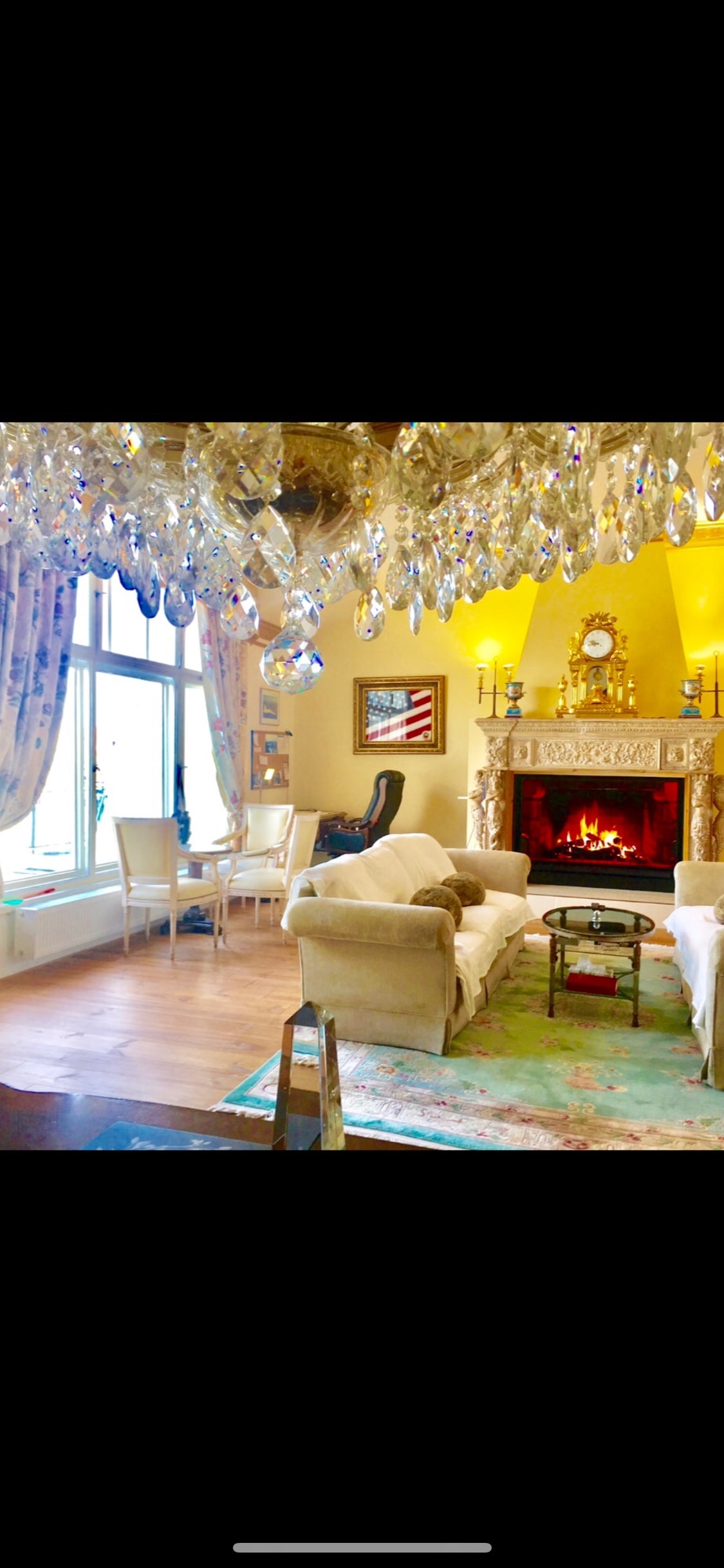 A spacious living room is illuminated by a sparkling chandelier, creating a warm ambiance. A fireplace is framed by decorative elements, while a comfortable sofa invites relaxation. Light streams through a large window, and a dining area with elegant chairs is positioned nearby.