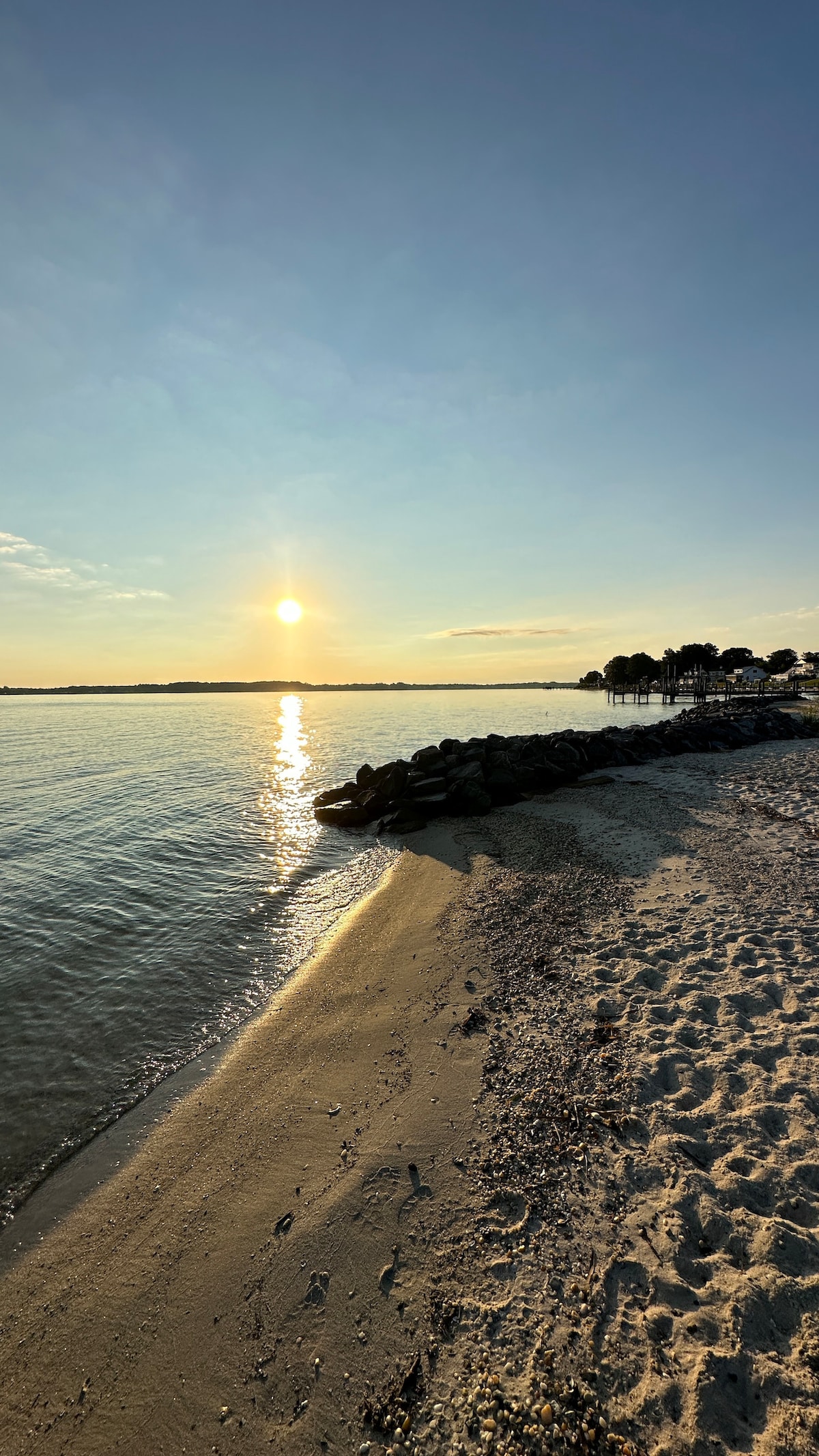 A serene waterfront scene captures the setting sun reflecting off calm water. Sandy shores curve along the coastline, with a rocky jetty visible on the left side. The sky exhibits soft hues, transitioning from blue to golden, while trees and structures line the distant shore.