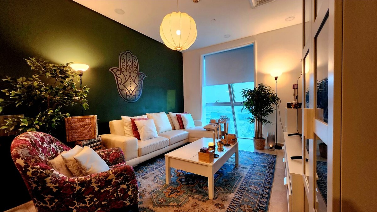 A modern living space features a cozy sofa and armchair, accented by decorative cushions. A large window offers a view of the water, and soft lighting from multiple sources illuminates the room. A decorative area rug lies beneath a central coffee table.