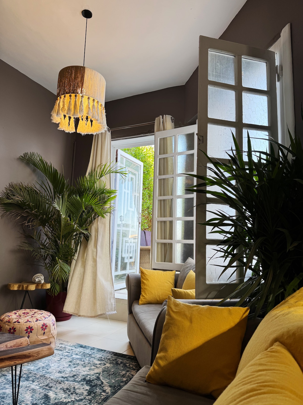 A comfortable sitting area is highlighted by large windows allowing natural light. Green plants add a fresh touch, while yellow cushions enhance the sofa's inviting look. A patterned rug rests underneath a low coffee table, offering a cozy spot to relax.
