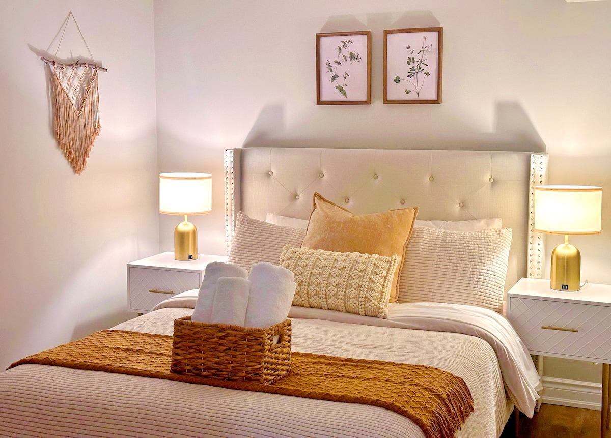 The bedroom features a spacious bed with a neutral-colored coverlet, flanked by matching nightstands topped with stylish lamps. Soft, light hues are complemented by decorative wall art and a woven wall hanging, creating a cozy and inviting sleeping area.