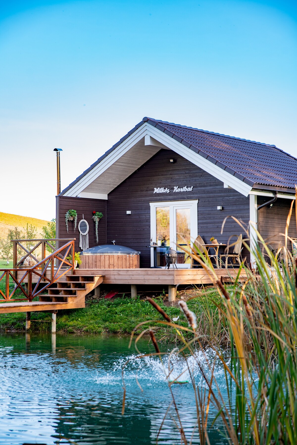 A newly built retreat is seen near a tranquil body of water, featuring a wooden deck with steps leading to the entrance. An outdoor hot tub is positioned on the deck, surrounded by lush greenery and reeds. Large glass doors provide access to the interior.