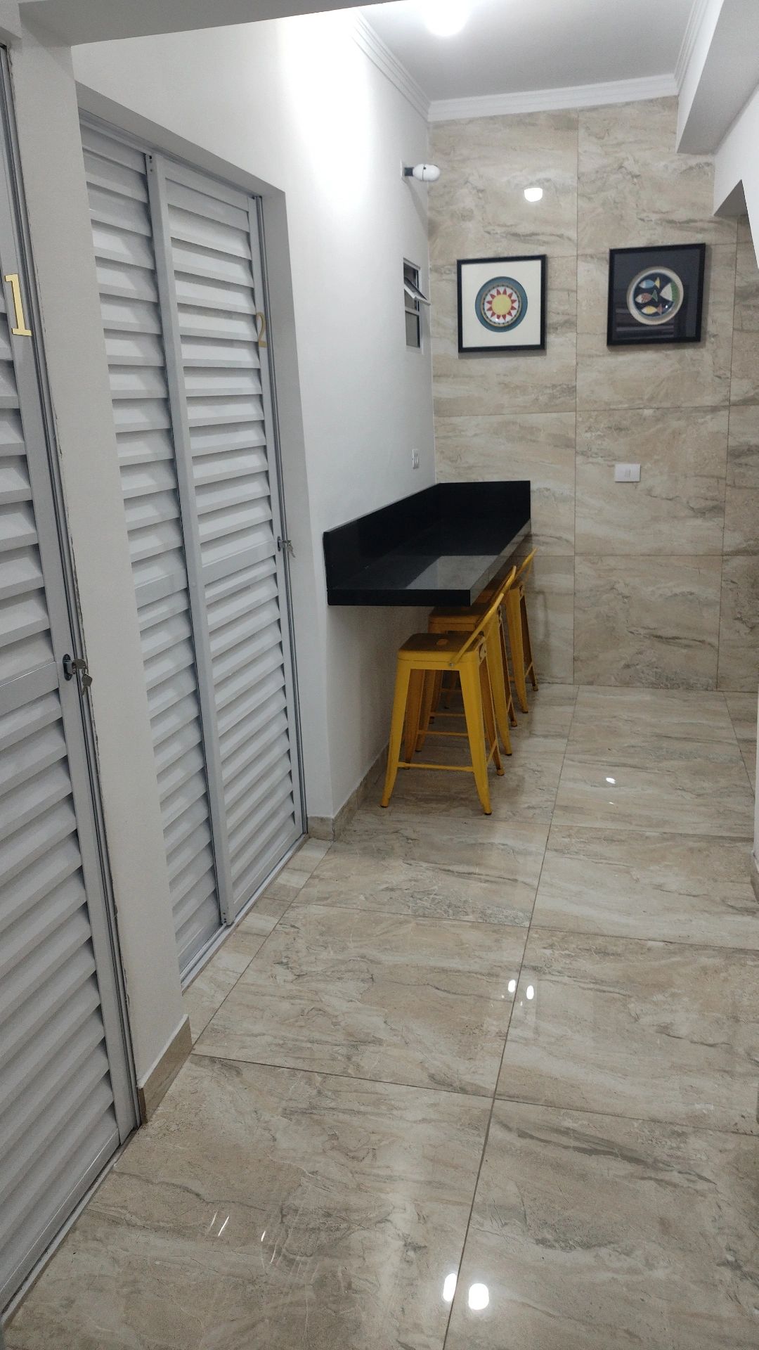 A narrow hallway is lined with smooth, tiled flooring, featuring a sleek black countertop against the wall. Two yellow stools are positioned beneath the counter, with framed artwork adorning the wall, complemented by white and gray tones of the surrounding space.