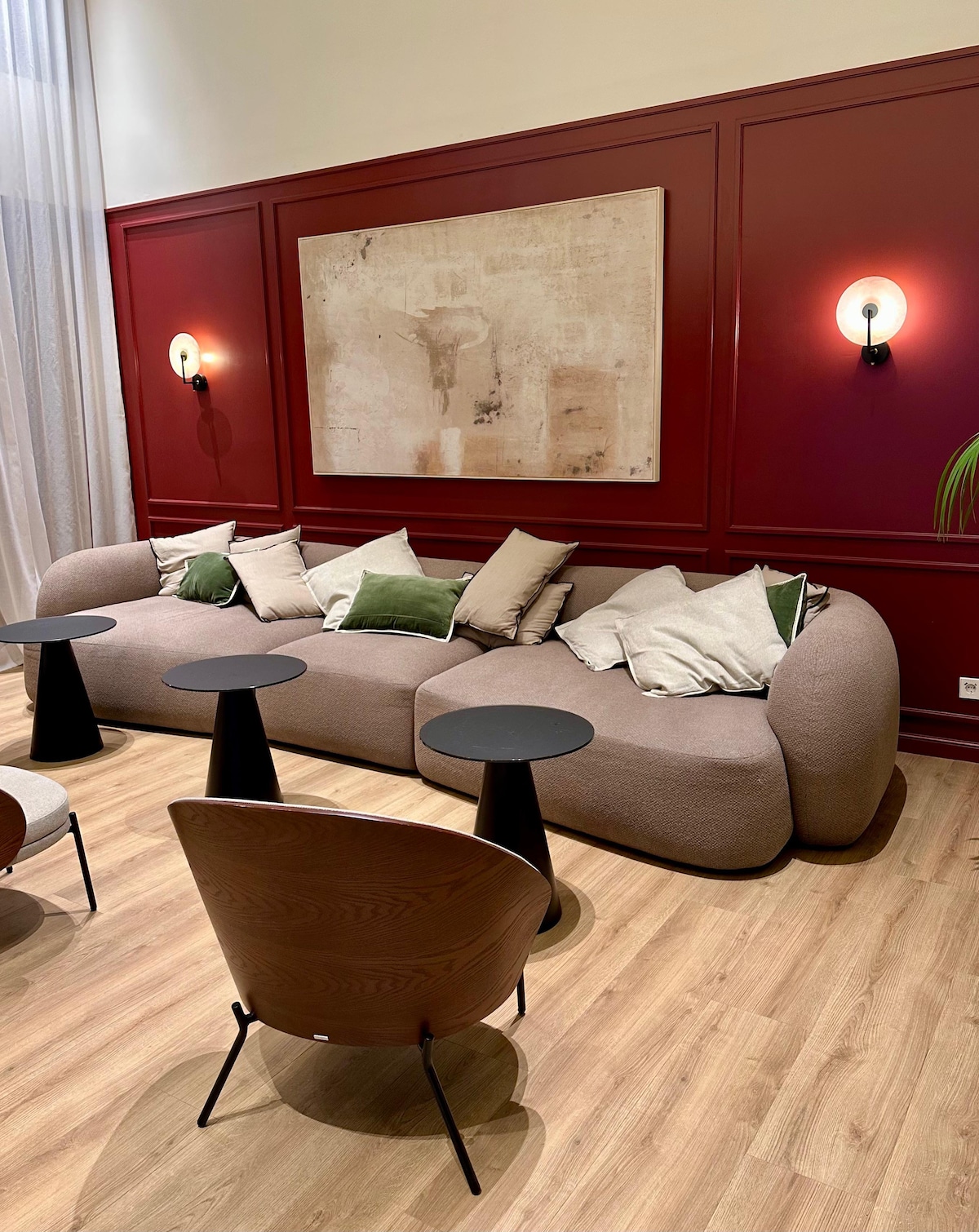 A stylish living area is presented, featuring a rounded, textured sofa adorned with multiple cushions in soft tones. Two sleek black tables complement the space, alongside minimalist wooden chairs. The backdrop is characterized by rich maroon walls, enhancing the modern aesthetic.