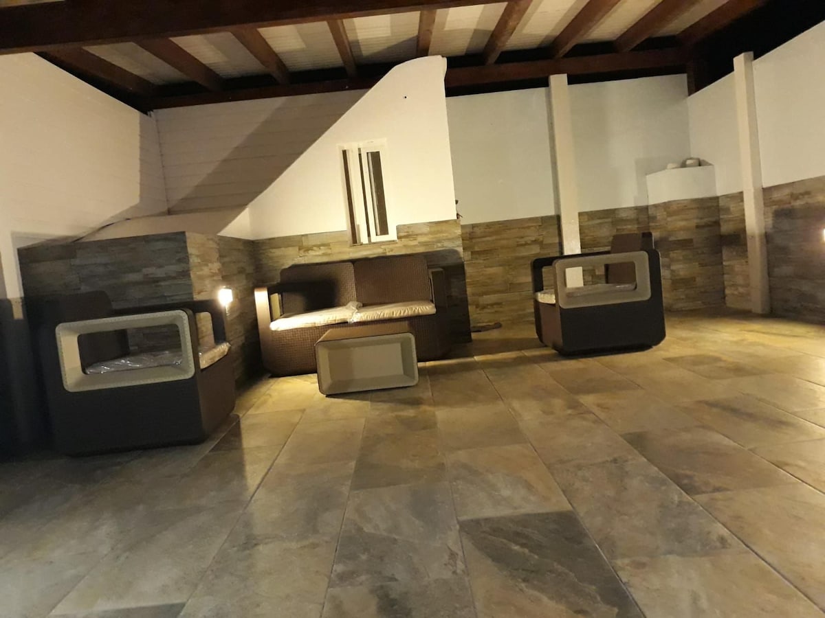 An outdoor seating area is presented, featuring modern lounge furniture arranged on tiled flooring. The space is illuminated with soft lighting, highlighting the stone walls and inviting a relaxed atmosphere. A partial view of a staircase is visible, adding to the area's openness.