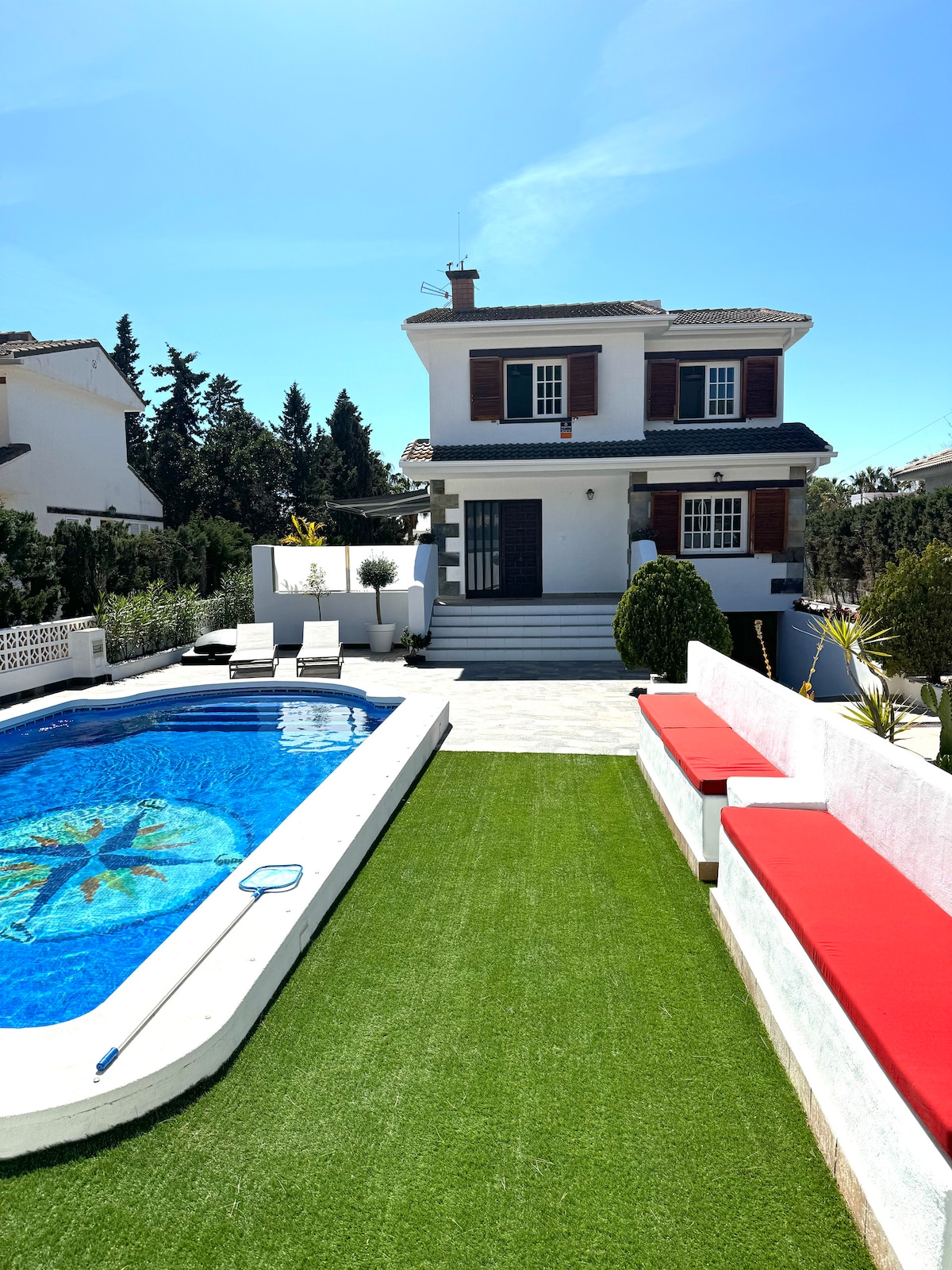 A two-story chalet is set amidst greenery, featuring a well-maintained pool with a decorative tile design. Lounge chairs are positioned around the pool, and a red cushioned seating area is visible adjacent to the lawn, providing ample outdoor relaxation space.