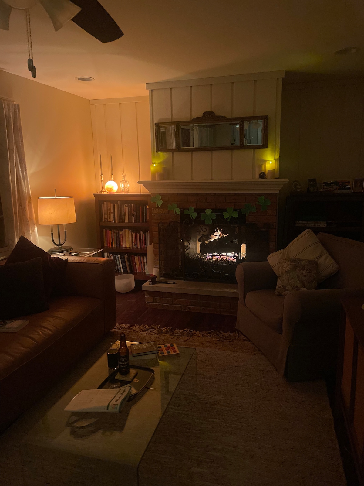 A cozy living room is illuminated by soft lighting, featuring a comfortable sofa and an armchair. A decorative fireplace is highlighted, with gentle flames visible through the glass. A wooden bookshelf filled with books adds to the inviting atmosphere, along with a coffee table displaying magazines.