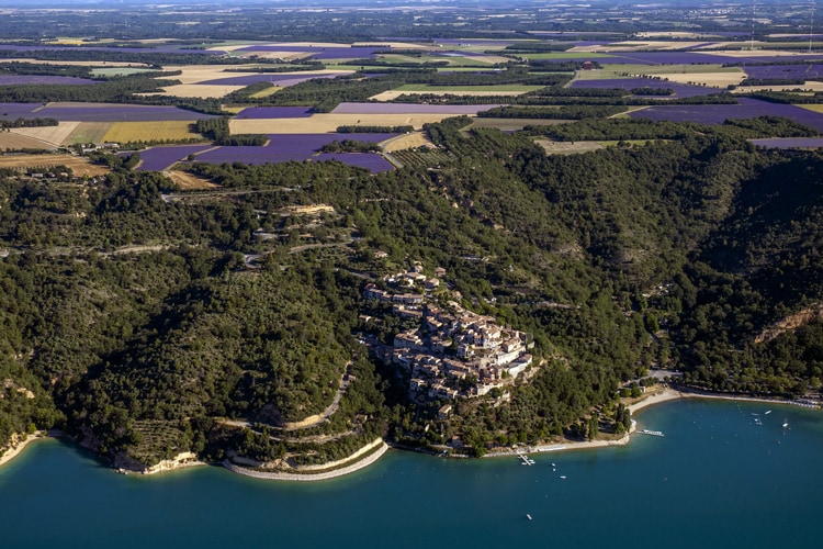 An aerial view captures the landscape surrounding Sainte-Croix-du-Verdon, showcasing green hillsides and the shimmering blue waters of the Lake. Fields of lavender are visible in the distance, complementing the scenic beauty of the calm waters and the charming village nestled among the trees.