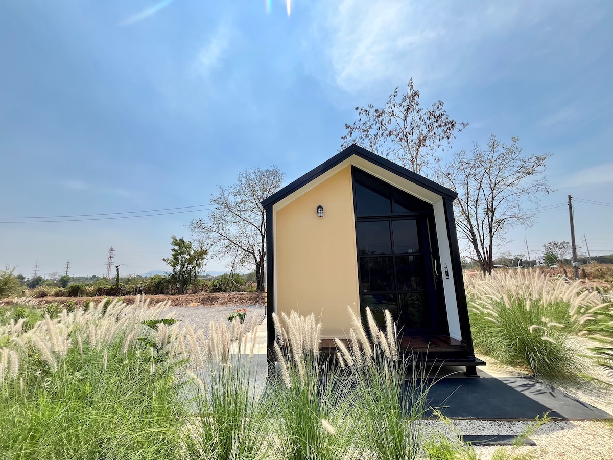 A modern, angular structure is surrounded by tall grass and shrubs. Large glass windows allow natural light to enter the interior. The building features a wooden entrance step and is set against a clear blue sky, with sparse trees in the background.