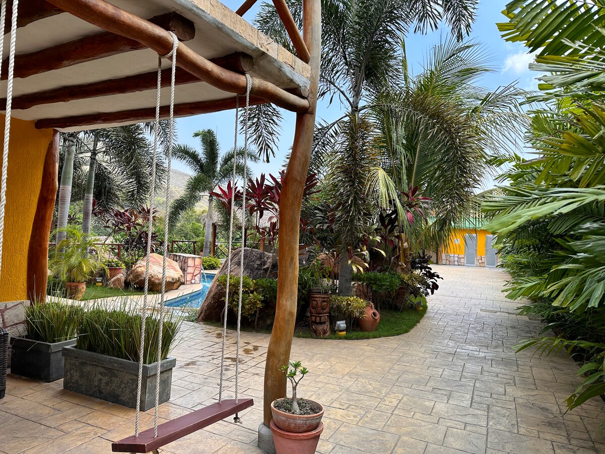 A wooden swing hangs from a tree, positioned in a lush garden area. Tropical plants and decorative pots surround the swing, creating a serene outdoor space. In the background, a swimming pool and additional buildings are subtly visible, enhancing the inviting atmosphere.