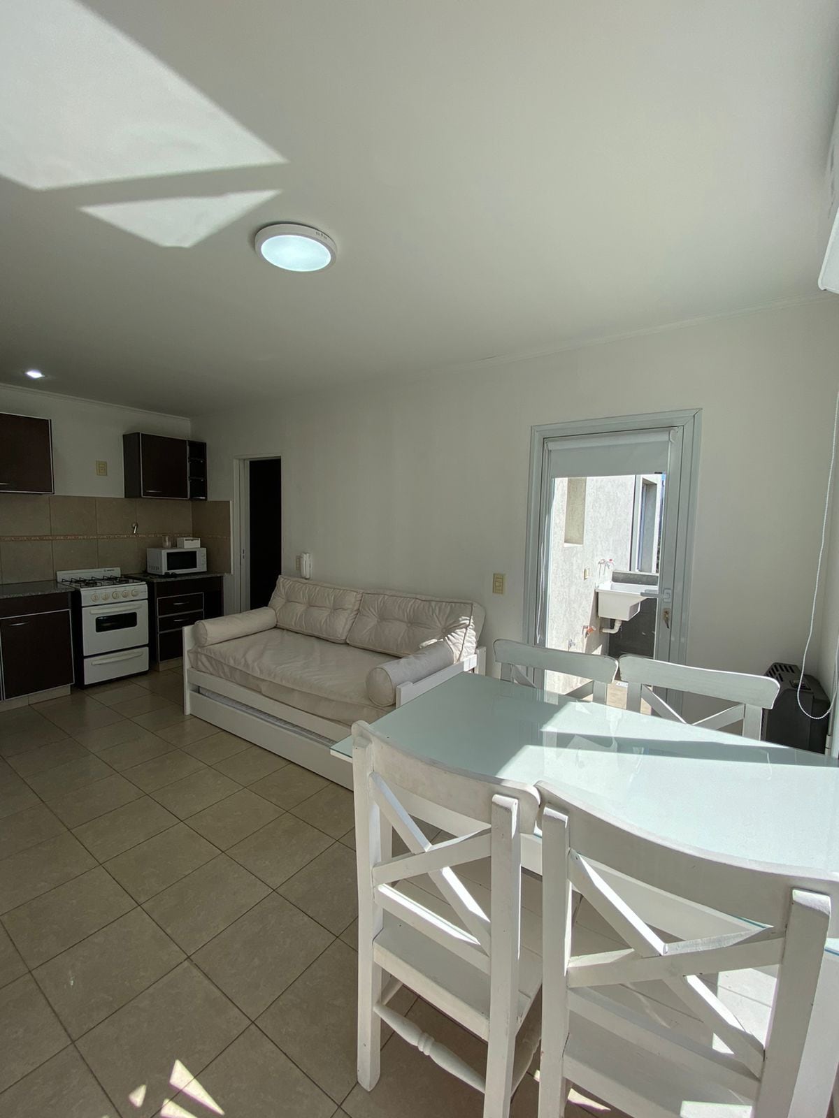 A bright living area is presented, featuring a comfortable light-colored sofa and a dining set with white chairs around a glass-topped table. The kitchen area is equipped with essential appliances, and a door leads to a balcony that allows natural light to filter in.
