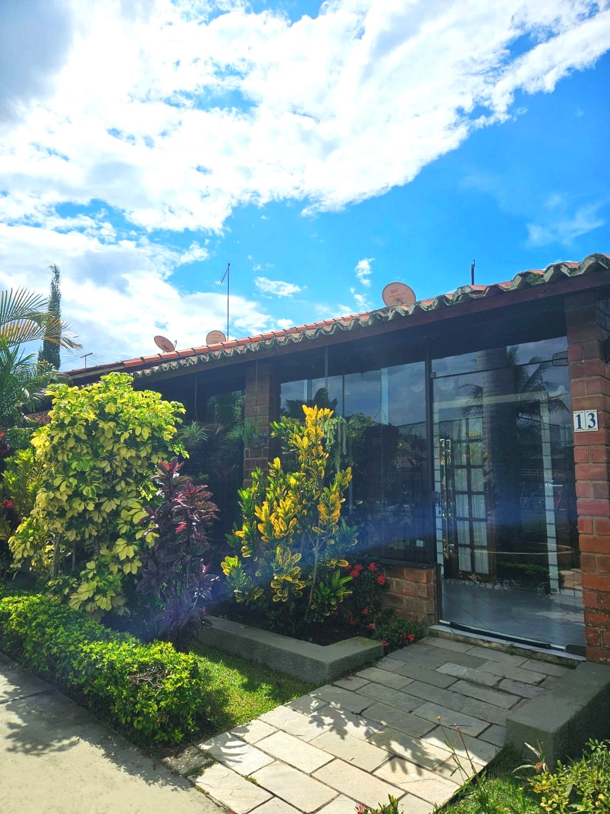 The exterior of the house features a combination of brick and glass walls, surrounded by a variety of colorful plants and flowers. A clear blue sky with scattered clouds accents the scene, creating a bright and inviting entrance.