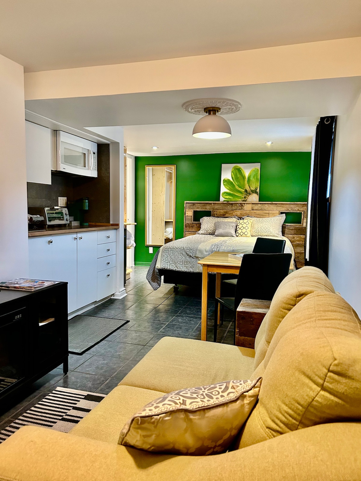 A compact studio is presented, featuring a living area with a comfortable sofa and a kitchenette in the background. The space is accented by vibrant green walls and includes a sleeping area with a spacious bed. Natural light is enhanced by overhead lighting and wall-mounted fixtures.
