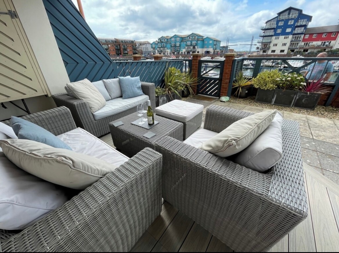 A stylish outdoor seating area is presented with comfortable woven furniture, featuring soft cushions and a small coffee table. The terrace overlooks the marina, offering views of boats and waterfront buildings under a partly cloudy sky.