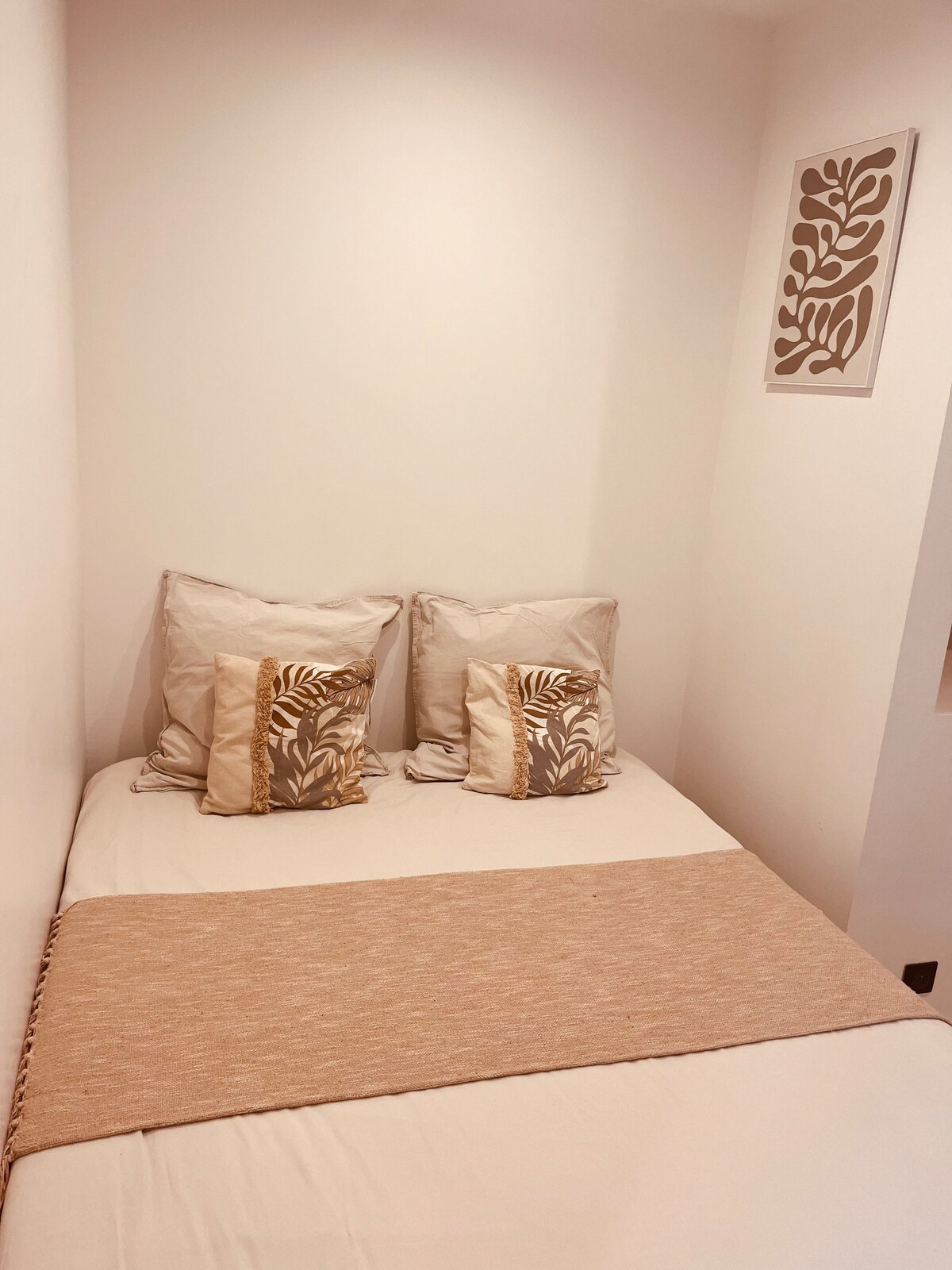 A comfortable sleeping area features a spacious bed with neutral-toned bedding and decorative pillows. A soft beige throw lies across the foot of the bed. A simple piece of wall art adds a touch of decor to the light-colored walls.